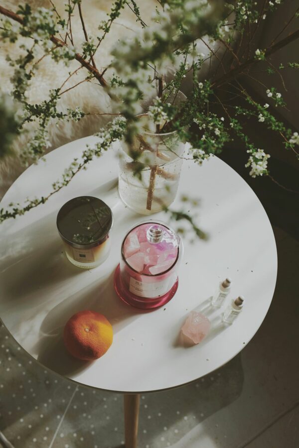 Elegant round table styled with luxury candles, a jar of rose quartz, perfume vials, a grapefruit, and a vase of delicate white blossoms, evoking a refined, botanical aesthetic.